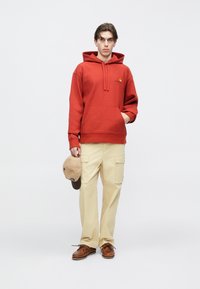 Κόκκινο φούτερ με κουκούλα και μπροστινή τσέπη, λογότυπο στο στήθος, σετ με μπεζ παντελόνια cargo και καφέ παπούτσια τύπου boat, κρατώντας ένα καπέλο σε ταμπά χρώμα.