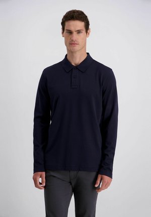 Mann mit kurzem lockigem Haar, der ein dunkelblaues, langärmeliges Poloshirt und dunkle Hosen trägt, steht vor einem schlichten hellen Hintergrund.