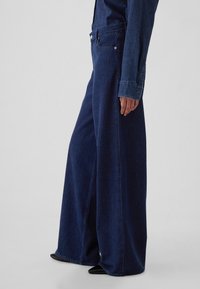 Sötétkék, széles szárú farmernadrág, sima textúrával, magas derékkal és elülső zsebekkel; egy hozzá illő farmeringgel kiegészítve.