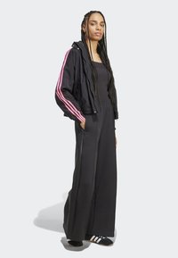 Musta kevyt vetoketjulla varustettu takki, jossa on vaaleanpunaisia raitoja, yhdistettynä mustiin leveälahkeisiin housuihin. Mallilla on päällään mustavalkoiset raidalliset liukujalkineet.