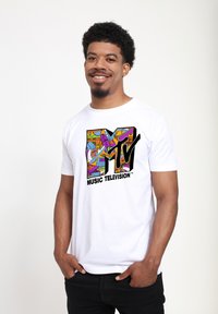 White cotton t-shirt featuring a colorful MTV logo with intricate designs. Model is wearing dark jeans, standing against a plain background.