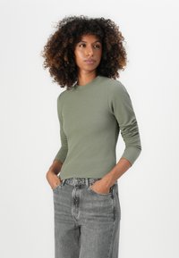 Olijfgroen geribbeld langslijfig shirt met een ronde halslijn, gecombineerd met hoge verwasemde grijze jeans, handen in de zakken, neutrale achtergrond.