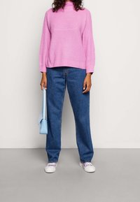 Kurzärmeliges, hellrosa gestricktes Pullover mit hohem Ausschnitt, kombiniert mit blauen Jeans mit geradem Bein und weißen Sneakern. Blaue Umhängetasche.