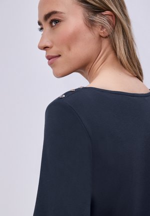 Vaaleahiuksinen nainen, jolla on tummansininen pusero, jossa on napit olkapäällä, katsoo sivulle vaaleaa taustaa vasten.