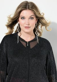Giacca nera con zip realizzata in materiale luccicante con scollatura trasparente. Presenta maniche ampie e orecchini decorativi.