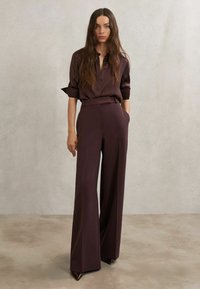 Tumši vīnsarkanā krekla ar uzpīpētām piedurknēm un pogu aizdari, kopā ar platām biksēm un smailiem papēžiem, attēlots uz neitrāla fona.