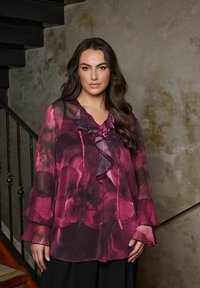 Blusa trasparente a maniche lunghe in tonalità di rosa intenso con dettagli a volant, motivo floreale e polsini a campana, abbinata a una gonna scura.