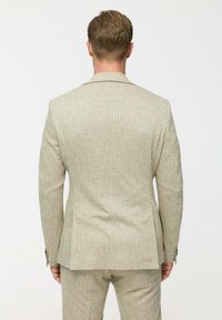 Férfi háttal áll, bézs mintás zakóval és hozzáillő nadrággal, sima fehér háttér előtt.