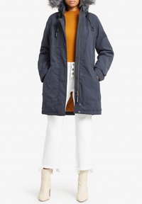 Navy parka jacket with a fur-lined hood, front zipper, two side pockets, and an inner orange lining, worn over a mustard turtleneck and white pants.