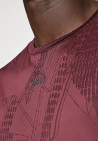 Burgunderfarbenes Sportshirt mit strukturiertem geometrischem Muster und dezentem Branding. Verfügt über einen runden Halsausschnitt und glattes, synthetisches Material.