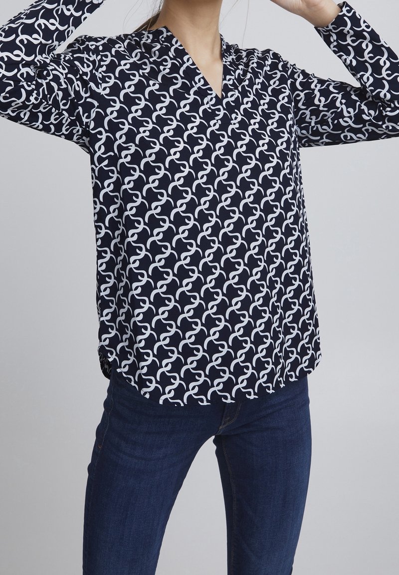 Navy blouse with white abstract pattern, long sleeves, and a V-neckline. Soft fabric with a relaxed fit and curved hemline.