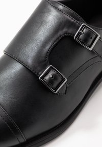 Chaussure de cuir noir avec une texture lisse, bout rond, boucles décoratives et détails cousus. La semelle est plate et légèrement courbée.