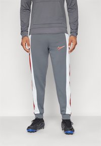 Hombre vistiendo joggers grises de Nike con franjas laterales blancas y rojas, botas negras con suelas azules, y una camiseta de manga larga gris, de pie sobre un suelo blanco.