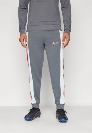 Mann trägt graue Nike-Jogginghosen mit weißen und roten Seitenstreifen, schwarze Schuhspikes mit blauen Sohlen und ein graues Langarmshirt, steht auf einem weißen Boden.