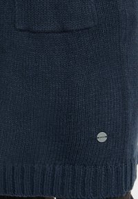 Marinblå stickad tröja med ribbat avslut, som har en framficka och en liten silverfärgad logotyp. Tillverkad av mjukt, texturerat garn.