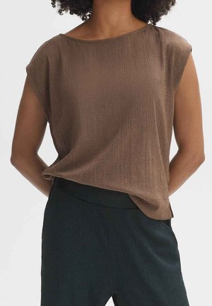 Donna che indossa un top senza maniche marrone a trama ampia infilato in pantaloni neri a trama alta in vita, su uno sfondo neutro.