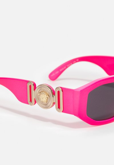 Rosa oversized solbriller med matte rammer, rektangulær form, mørke linser, og gull sirkulært emblem med løve-detall på stengene.