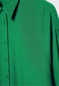 Chemise verte à boutons, tissu lisse, col et manches longues. Texture notable avec un motif de tissage régulier.