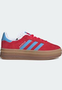 Röda mockasneakers med blå ränder, rund tå, kraftig gummisula och vit texturerad krage. Har "Gazelle"-märkning på sidan.