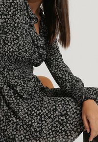 Vestido negro con estampado floral blanco, escote con volantes, mangas largas y cintura fruncida. Tejido suave con una silueta fluida.