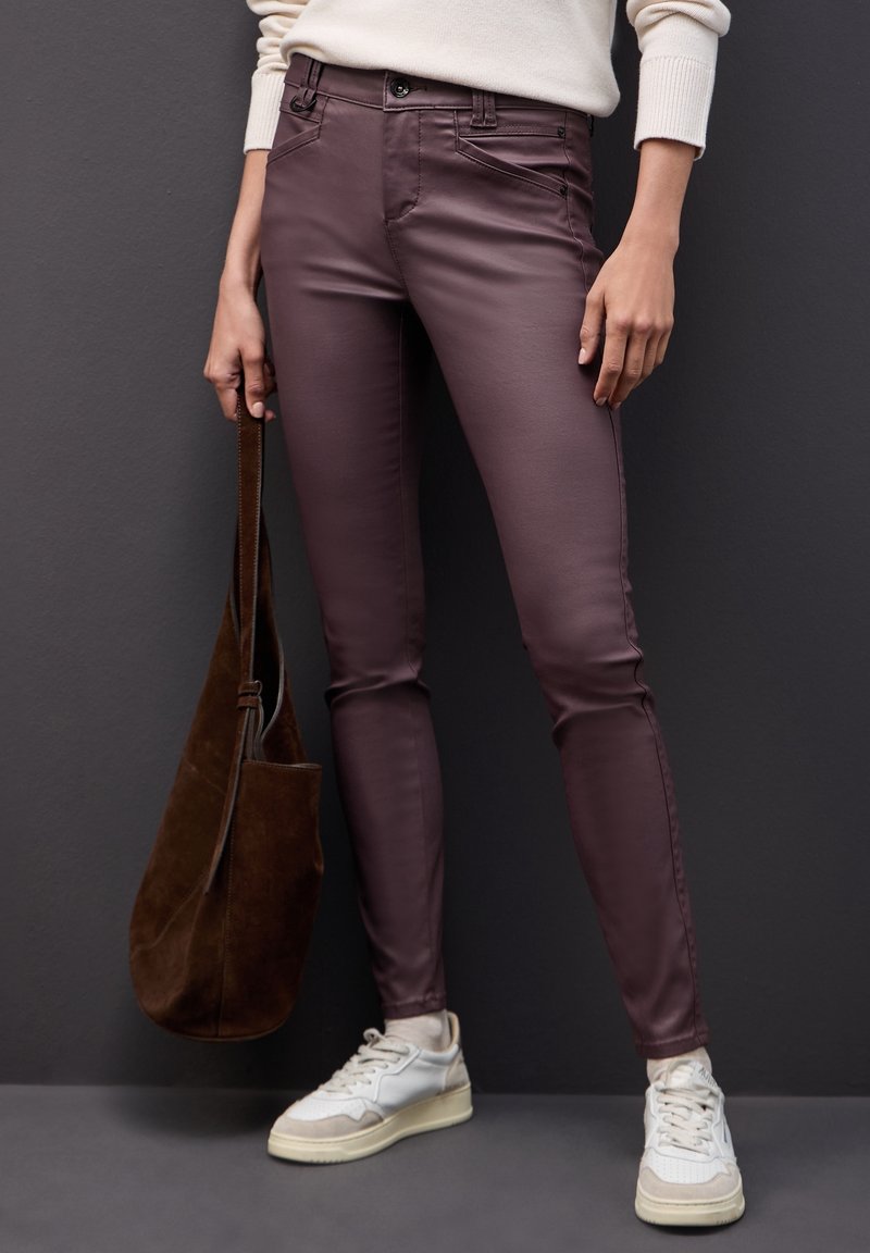Personne portant un pantalon slim violet foncé, des baskets blanches et un pull crème, tenant un grand sac en daim marron sur fond sombre.