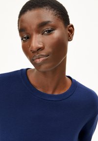 Pull bleu marine avec un col rond et un tissu texturé, porté par une personne avec des cheveux courts et foncés, sur un fond neutre.