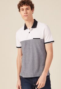Polo con un diseño de bloques de color blanco y gris, hecho de tejido texturizado, con un cuello y un bolsillo negros. Mangas cortas y detalles en contraste.