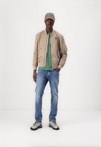 Beige bomberjakke med ribbede mansjetter, grønn t-skjorte, denimjeans og sølvfargede joggesko. Capsen har tekst med "Worn". Enkelt design uten mønstre.