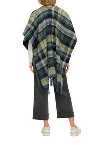 Geruiten poncho in donkere groentinten, grijs en geel met franjes aan de randen. Losse, gedrapeerde stijl, gedragen over een witte top en grijze broek.