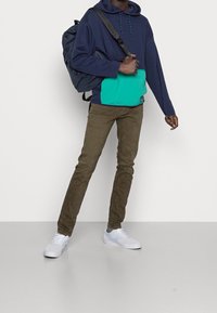 Person klädd i en marinblå huvtröja med tealfärgad ficka, bruna byxor, vita sneakers och bärande en marinblå ryggsäck över ena axeln.