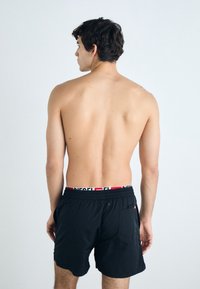 Black swim shorts with an elastic waistband featuring a red and white logo. Smooth texture, side pockets, and a back pocket detail.