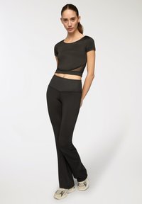 Maglietta nera corta con maniche corte e dettagli trasparenti, abbinata a leggings neri attillati. Il modello indossa scarpe sportive beige.