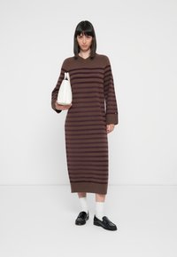 Καφέ και μωβ ριγέ πλεκτό φόρεμα με v-λαιμόκοψη, μακριά μανίκια και ίσια σιλουέτα, συνδυασμένο με λευκή τσάντα και μαύρα δερμάτινα παπούτσια.