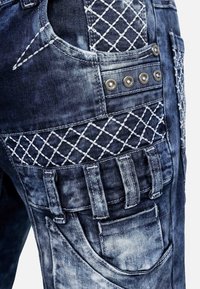 Jeans in denim con un lavaggio blu sbiadito, caratterizzati da cuciture incrociate bianche, accessori in argento e molteplici tasche sui lati.