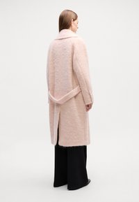 Casaco de lã rosa claro com uma silhueta descontraída, grande gola, mangas largas e detalhe de laço nas costas. Acabamento texturizado com fendas laterais.