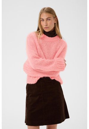 Mujer con un suéter rosa esponjoso sobre un cuello alto negro y una falda negra, de pie con los brazos cruzados frente a un fondo liso.