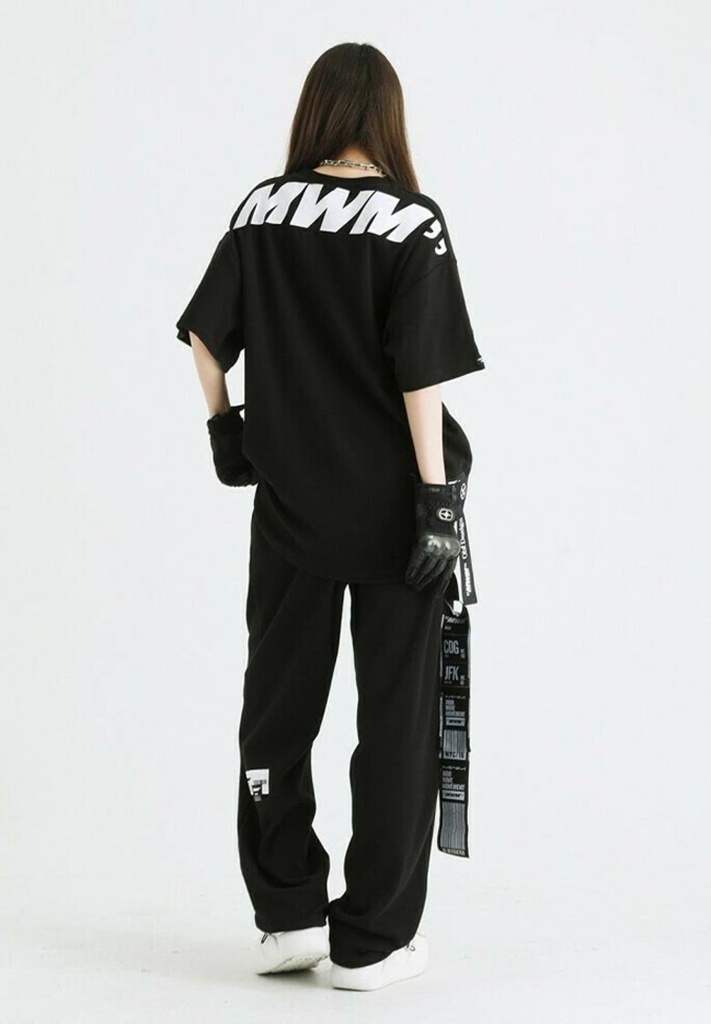 Camiseta negra de gran tamaño con letras blancas en la parte de atrás, combinada con pantalones negros que presentan un logo y tirantes de patrón, y guantes negros.