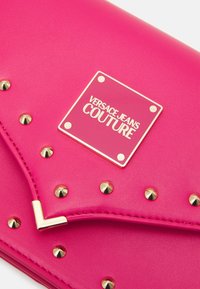 Clutch de color rosa neón con tachuelas doradas y placa con logo. Textura suave y diseño de solapa en ángulo. Presenta un acento metálico para un cierre seguro.