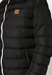 Chaqueta de plumas negra con acolchado horizontal, cremallera blanca y una etiqueta marrón "Urban Classics" en la parte superior izquierda. Tejido suave y brillante.