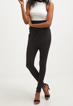 Mujer de pie con los brazos cruzados, vestida con una blusa sin mangas blanca y negra, pantalones ajustados negros y sandalias negras de tacón alto.