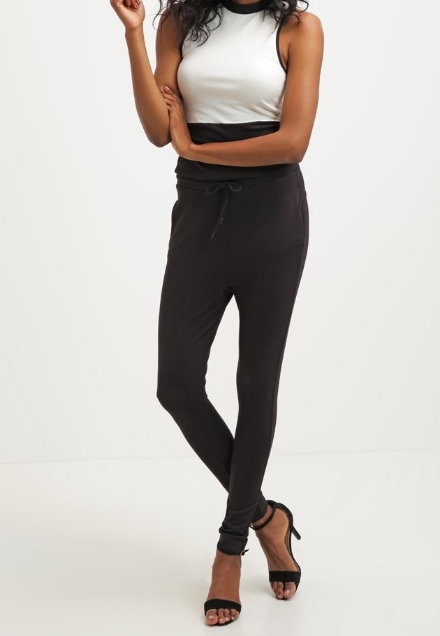 Femme debout, les bras croisés, portant un haut sans manches blanc et noir, un pantalon noir ajusté, et des sandales à talons hauts noires.