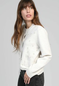 Femme aux longs cheveux bruns portant un cardigan blanc à boutons avec de petits détails brodés, un pantalon noir, regardant sur sa gauche.
