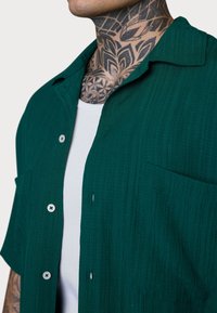 Nahaufnahme einer Person, die ein dunkelgrünes, strukturiertes Hemd mit Knöpfen über einem weißen Tank-Top trägt, wobei sie aufwendig gestaltete Nackentattoos zeigt.