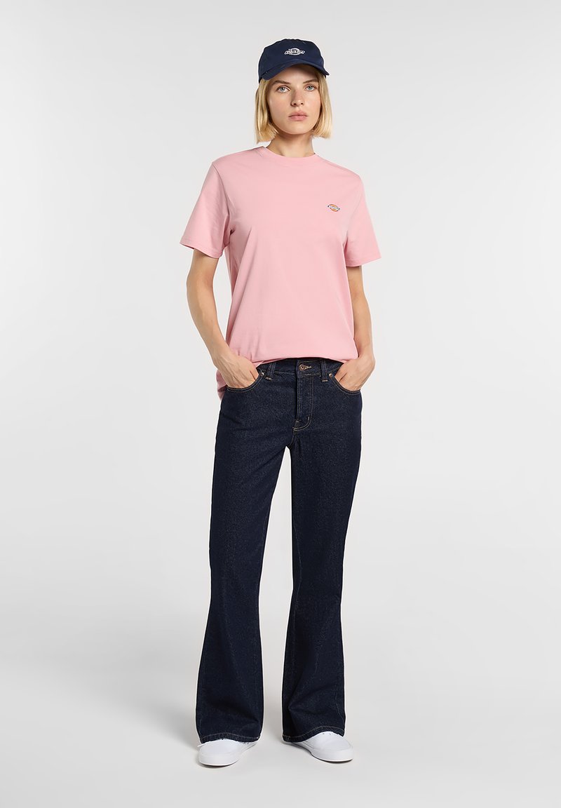Pink cotton t-shirt with a round neck, paired with dark blue flared denim jeans. White sneakers and a navy cap complete the look.