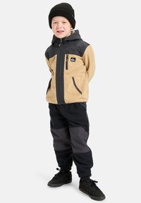 Chaqueta de forro polar beige y negra con bolsillos con cremallera, combinada con pantalones de forro polar negros y zapatillas negras. El modelo lleva un gorro de punto negro.