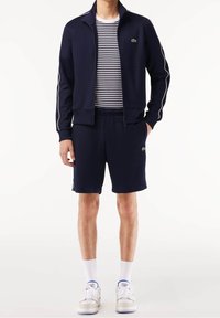 Homme portant une veste de survêtement bleu marine avec des passepoils blancs, un short assorti bleu marine, une chemise rayée noir et blanc, des chaussettes blanches et des baskets blanches.