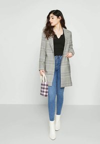 Blazer comprido cinza xadrez com um corte sob medida, blusa preta e jeans skinny azul claro. Botas brancas de tornozelo e uma pequena bolsa tecida roxa.
