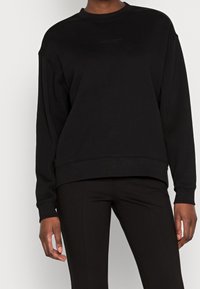Černá mikina s kulatým výstřihem a dlouhými rukávy, volný střih, texturovaná tkanina a minimální designové detaily. V kombinaci s černými upnutými kalhotami.