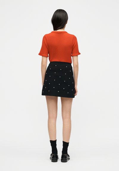 Γυναίκα που στέκεται γυρισμένη πίσω, φορώντας κόκκινη μπλούζα με κοντά μανίκια, μαύρη μίνι φούστα με λευκά σχέδια σταυρού, μαύρες κάλτσες και μαύρα παπούτσια.