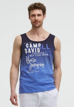 Mann mit braunen Haaren, der ein blaues ärmelloses Shirt mit weißem Text und weiße Hose trägt, steht mit einer Hand in der Tasche.